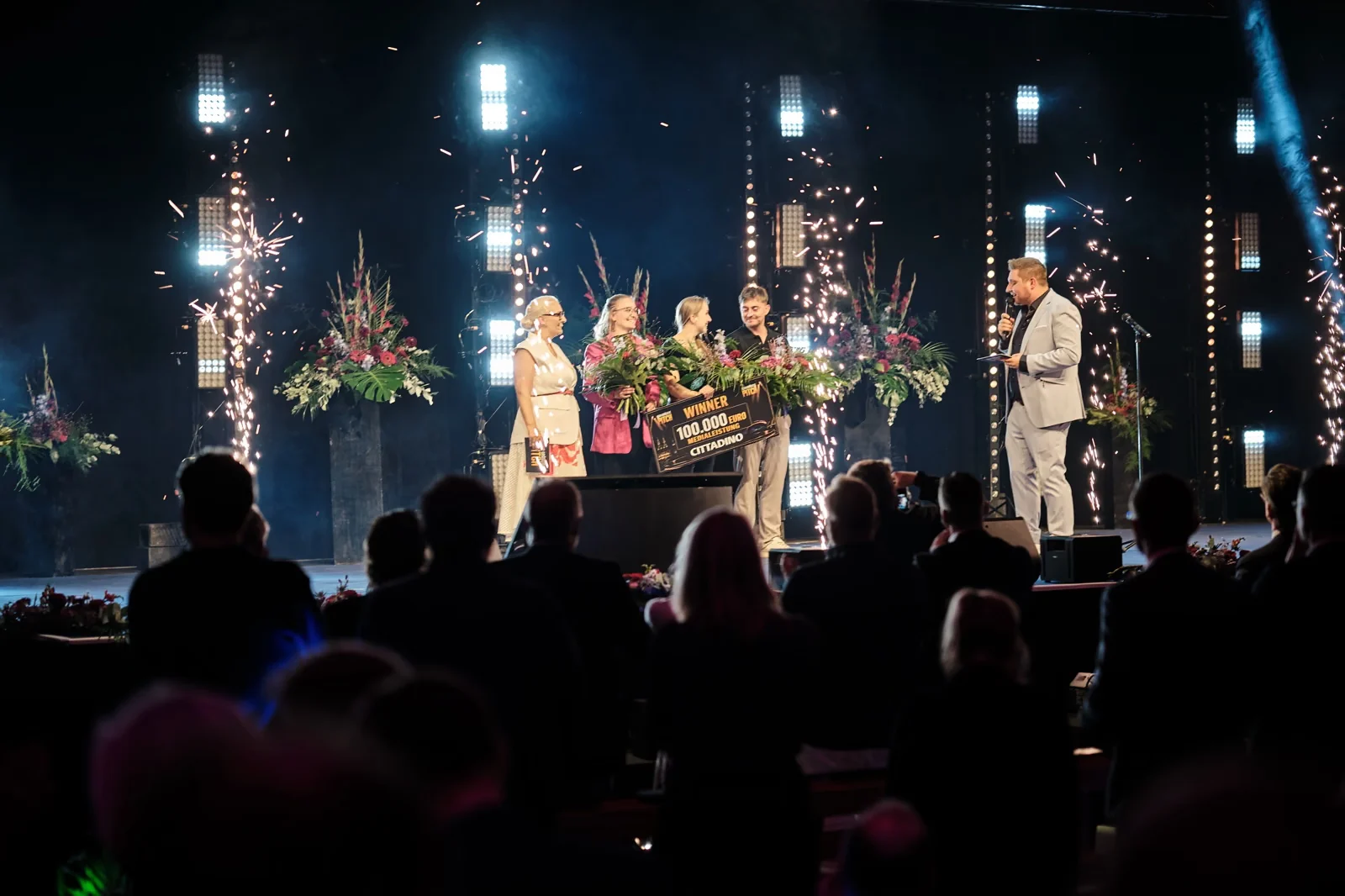 Siegerteam mit 100.000-Euro-Schild und Blumen auf der Arena-Bühne, Funkenfontänen im Hintergrund