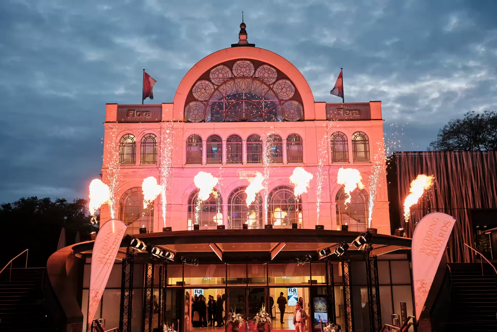 Kölner Flora bei Dämmerung, in Rosa angestrahlt, mit Bodenfeuer-Fontänen vor dem Eingang zu den Wedding King Awards