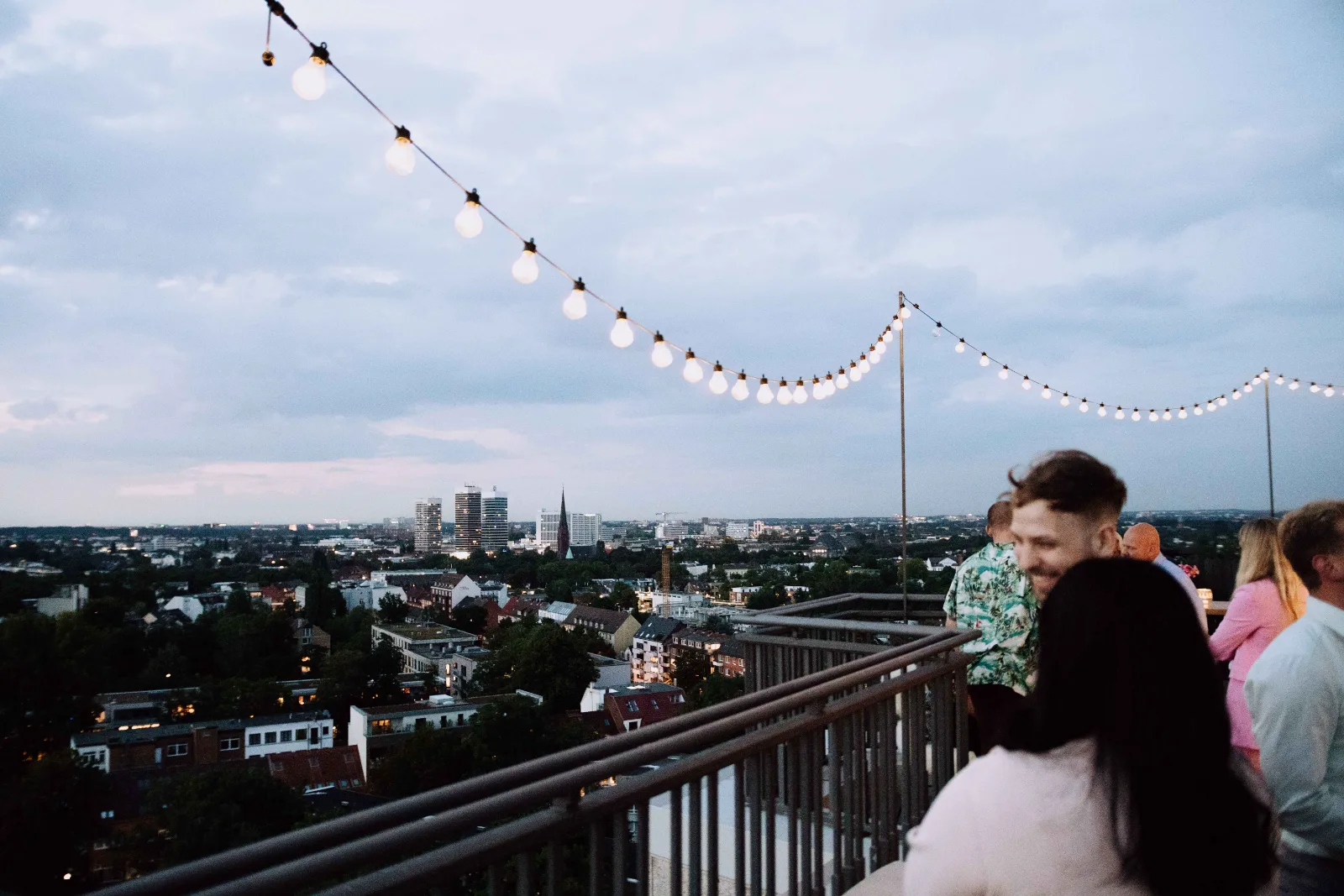 Gäste auf einer Dachterrasse unter Lichterketten mit Blick über die Stadt in der Abenddämmerung
