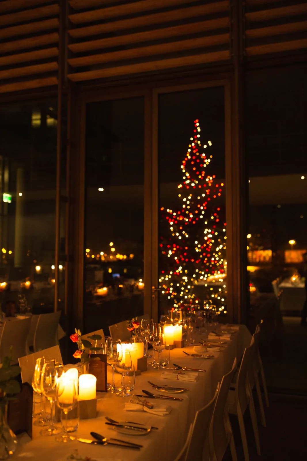 Festlich gedeckte Tafel mit Kerzen vor einem beleuchteten Weihnachtsbaum hinter großen Fenstern