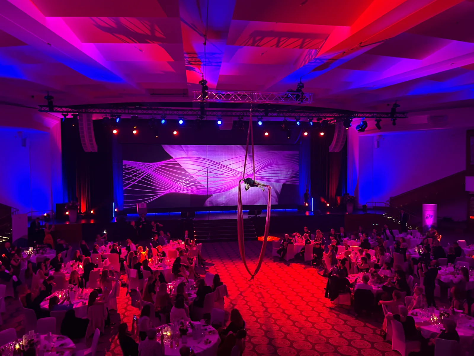 Gala-Abend mit Artistin an Vertikaltuch über der Tanzfläche, rot-blaues Licht und voll besetzte Tische